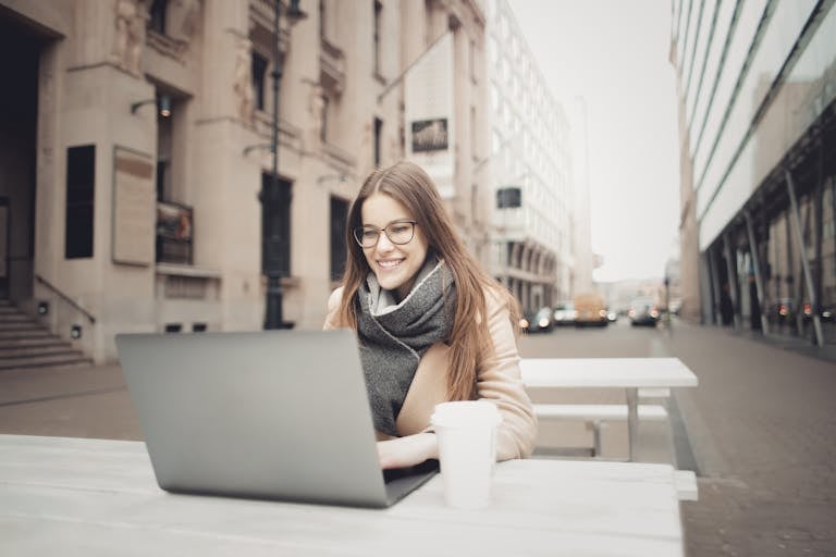 Young woman, which is a digital and remote worker, enjoying coffee while working on her laptop outdoors in a city in Italy.